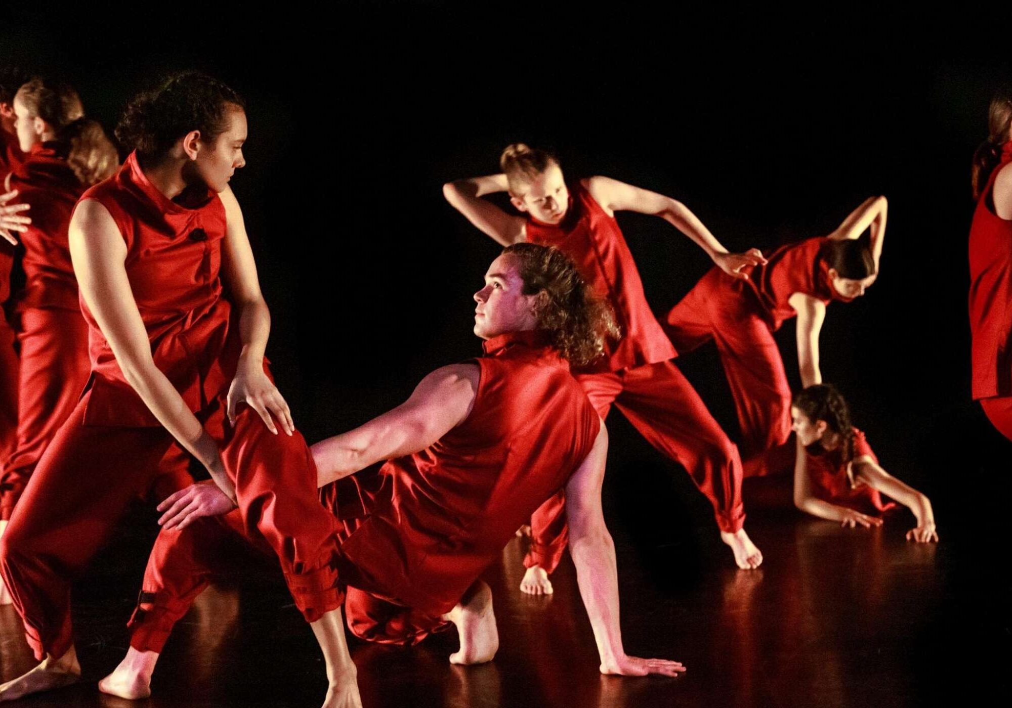 Seven dancers wearing loose, red tops and pants on a black stage, either standing or positioned on hands and knees