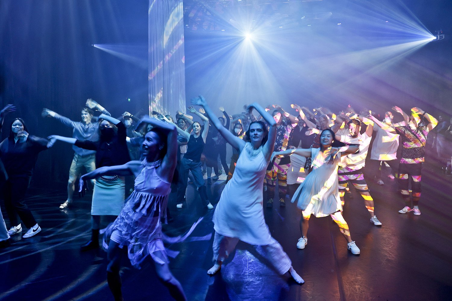 People of various ages and genders dancing together in a disco. Their arms are swaying in the air and bright laser lights shine across the room. 