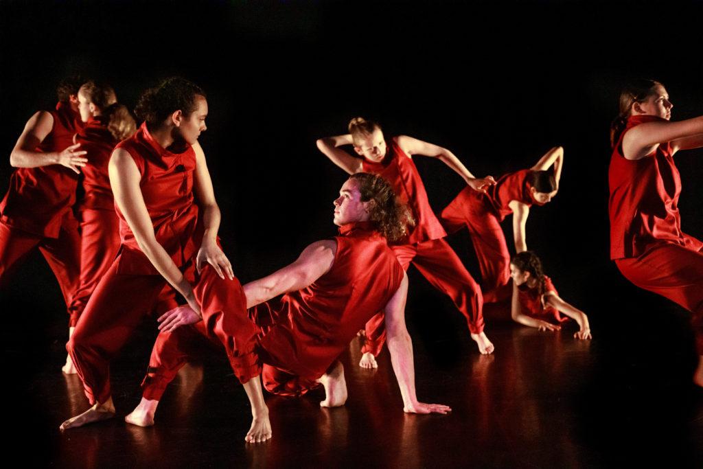 Seven dancers wearing loose, red tops and pants on a black stage, either standing or positioned on hands and knees