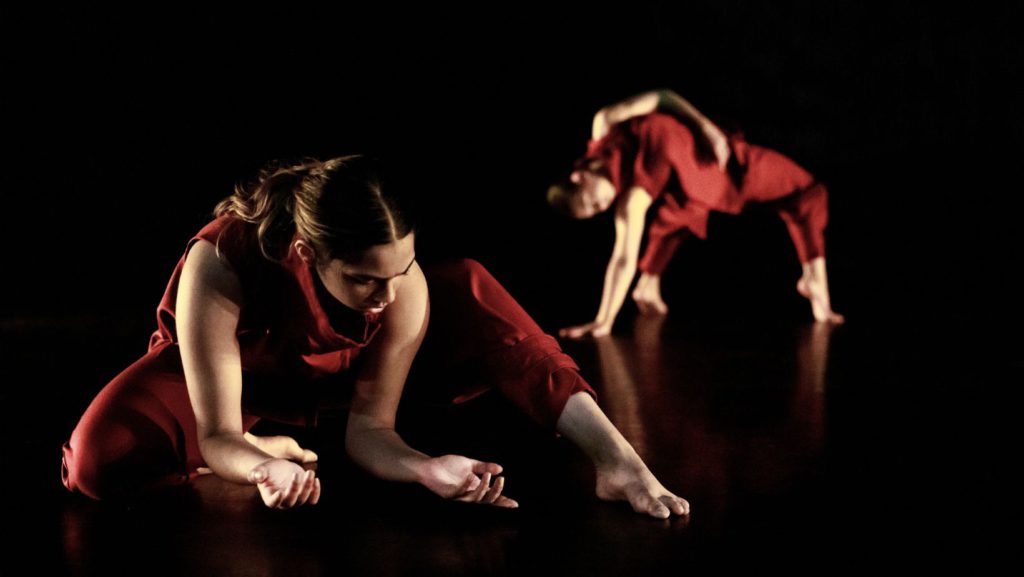 Image of a dance sitting on the ground with her palms facing up. In the background a dancer is bending over backwards with one arm wrapped around her body.