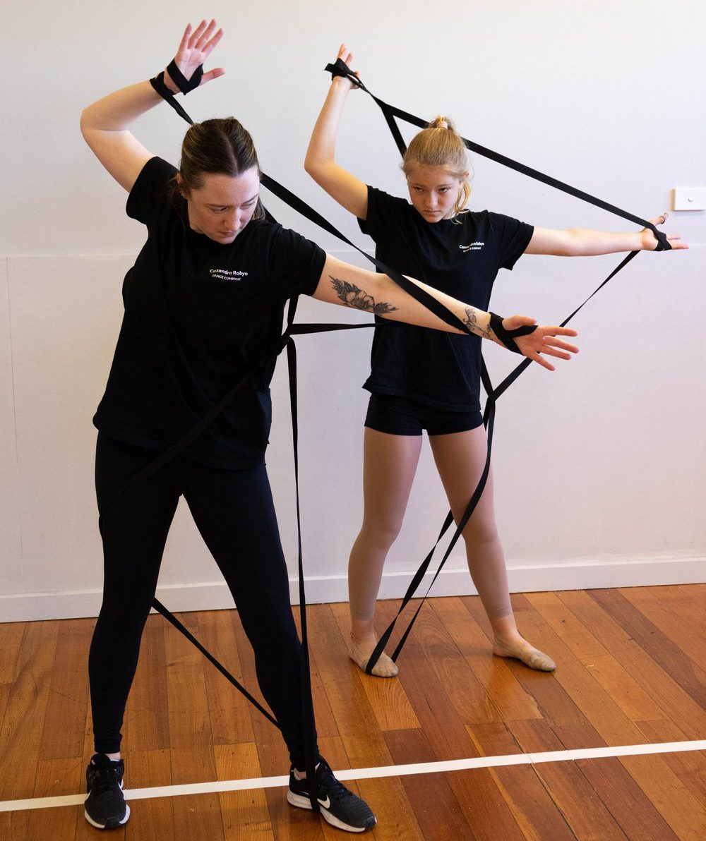 A dance teacher standing in a studio wearing black with black elastic winding and stretched around her leg, body, head and between both arms. A student is in the background mimicking the pose with black elastic.