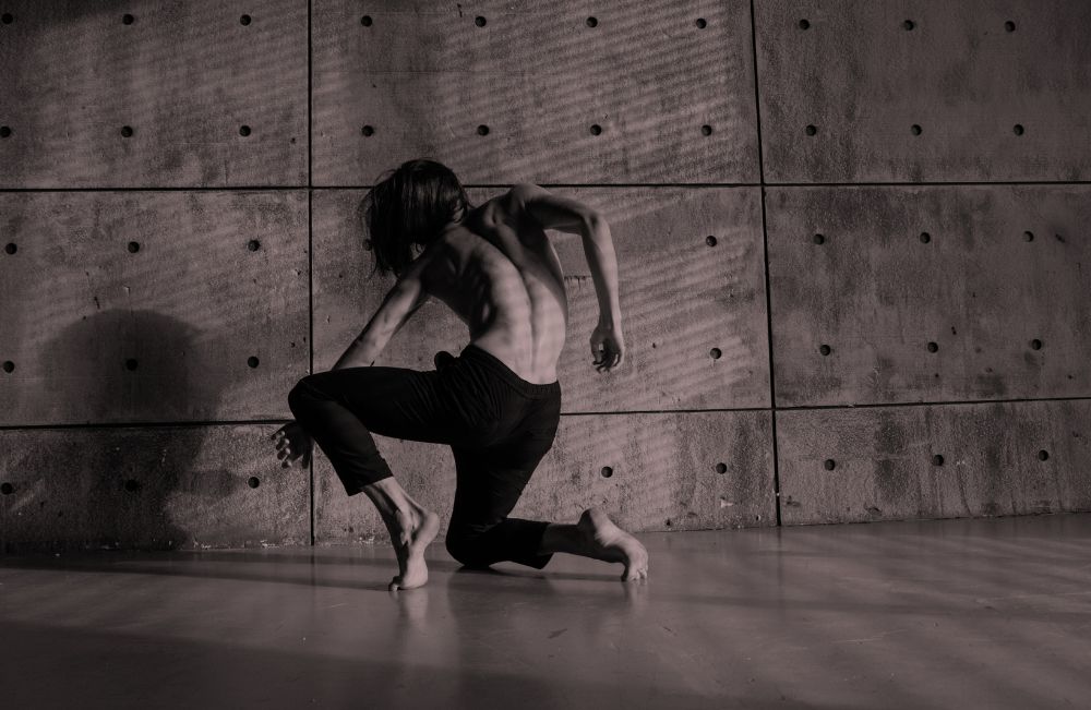A dancer wearing loose black pants kneels on the ground with their back to the viewer. They are against a concrete wall and floor.
