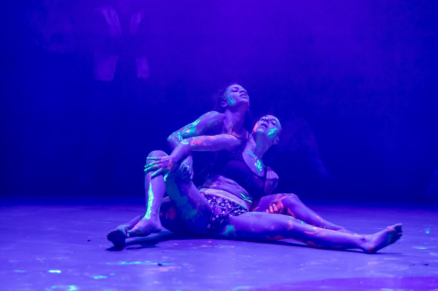 Two performers lay on the floor, holding each other. They are wearing underwear and their bodies are painted with neon paint in various colours. The lighting is low and dark.