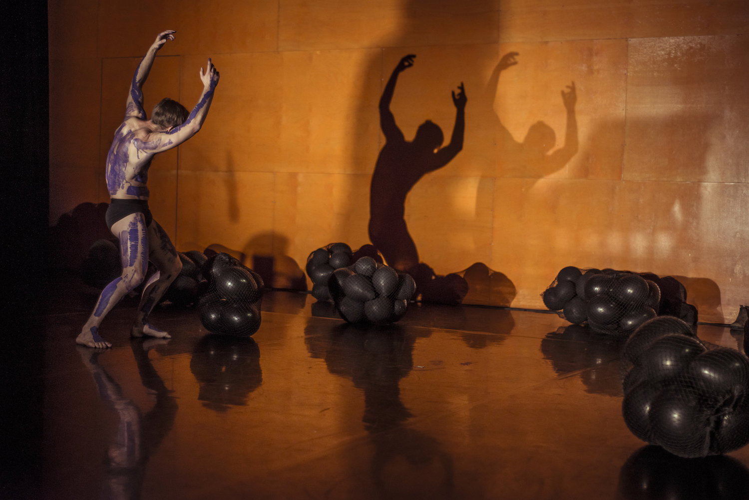 Joel Bray performing in Dharawungara. He is wearing small black trunks and his body is covered in large purple paint strokes. He is creating a shadow on the wall and there are groups of black balloons tied in nets around him on the floor.