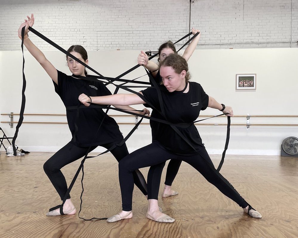 Three dancers in a studio wearing black with black elastic stretched and winding around their bodies. They are standing close together and using their outstretched arms and legs to create strong angles with their bodies.